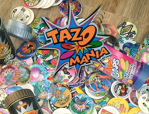 Foto principal do post, uma mesa de madeira cheia de tazos de varios tipos, porta tazos e a logo do TAZO mania escrita no meio